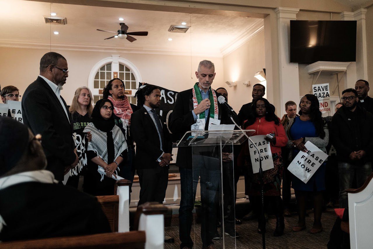 A group of individuals gathered in a church setting, with one person addressing a microphone while others stand around him holding signs of protest or awareness.