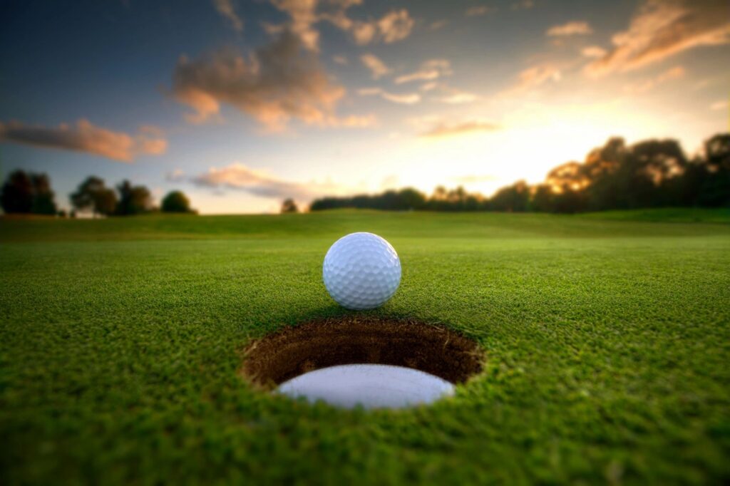 Golf ball on the edge of a hole on a green field at sunset, with a vibrant sky in the background.