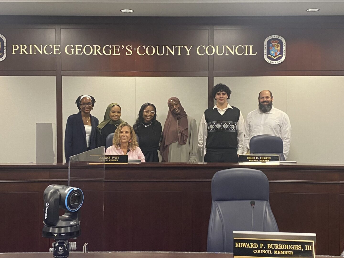 A group of seven people stand in front of the Prince George's County Council sign, including one seated at the desk. The desk has nameplates for council members. A camera is in the foreground.