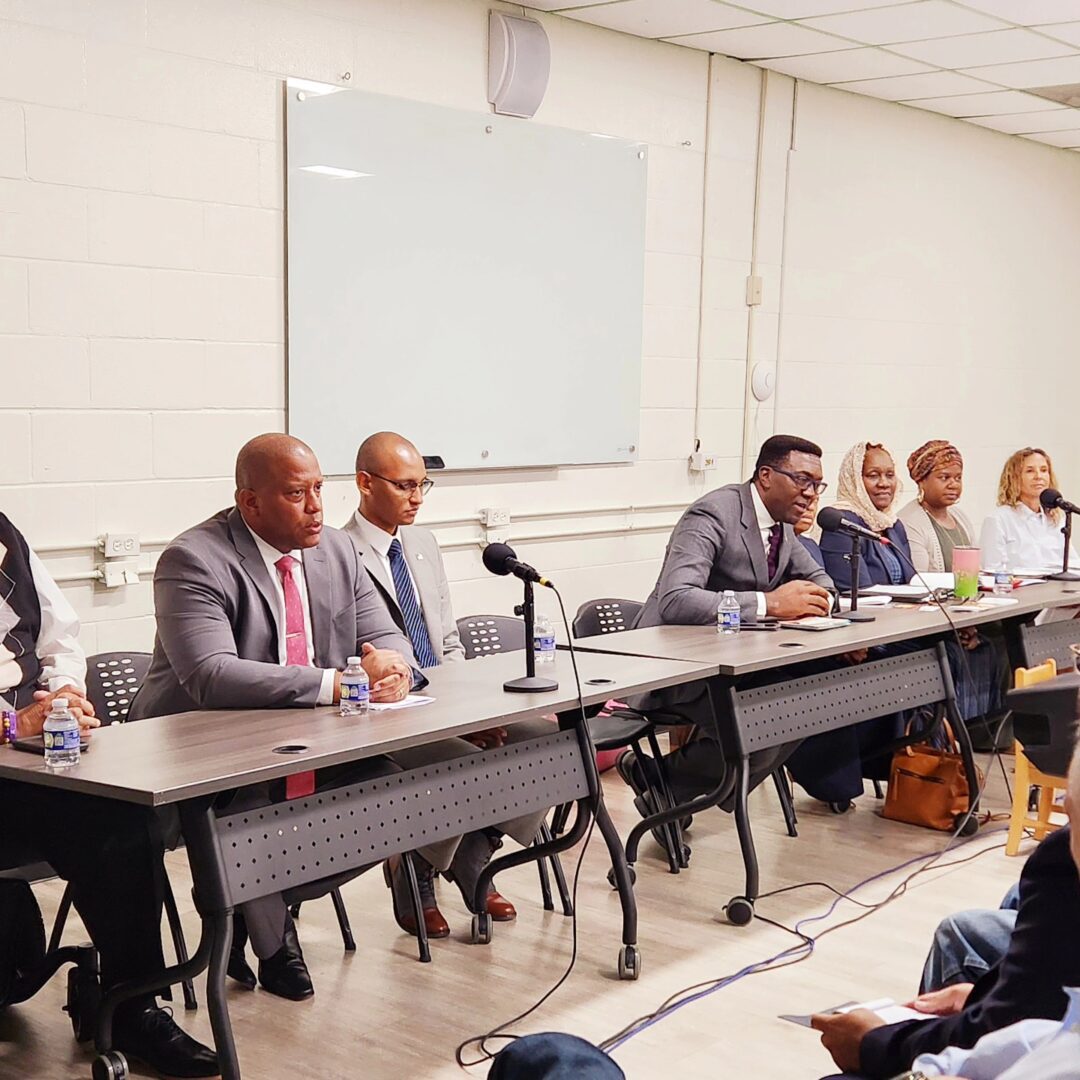 A group of people in formal attire sit at a long table with microphones, participating in a panel discussion in a room with a whiteboard on the wall.