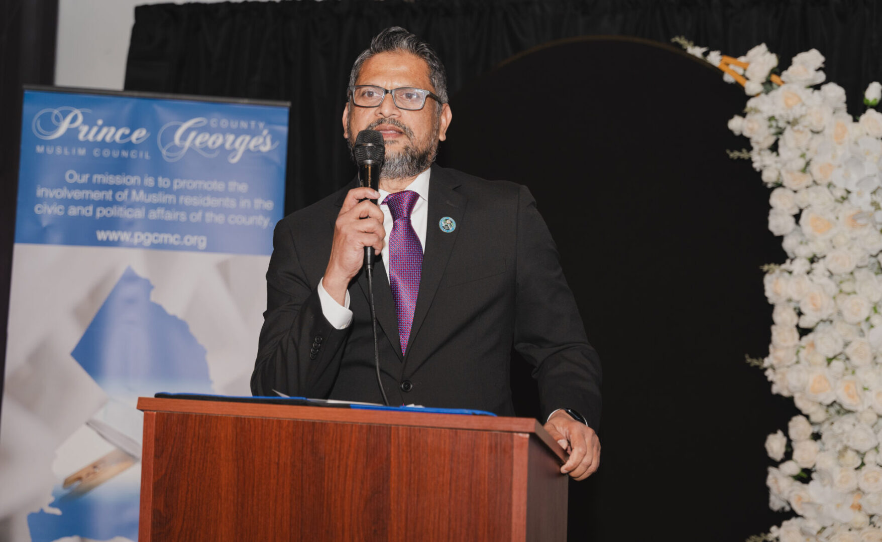 A man in a suit speaks into a microphone at a podium during an event. A sign on the left reads "Prince George's County Muslim Council." There's a floral decoration in the background.