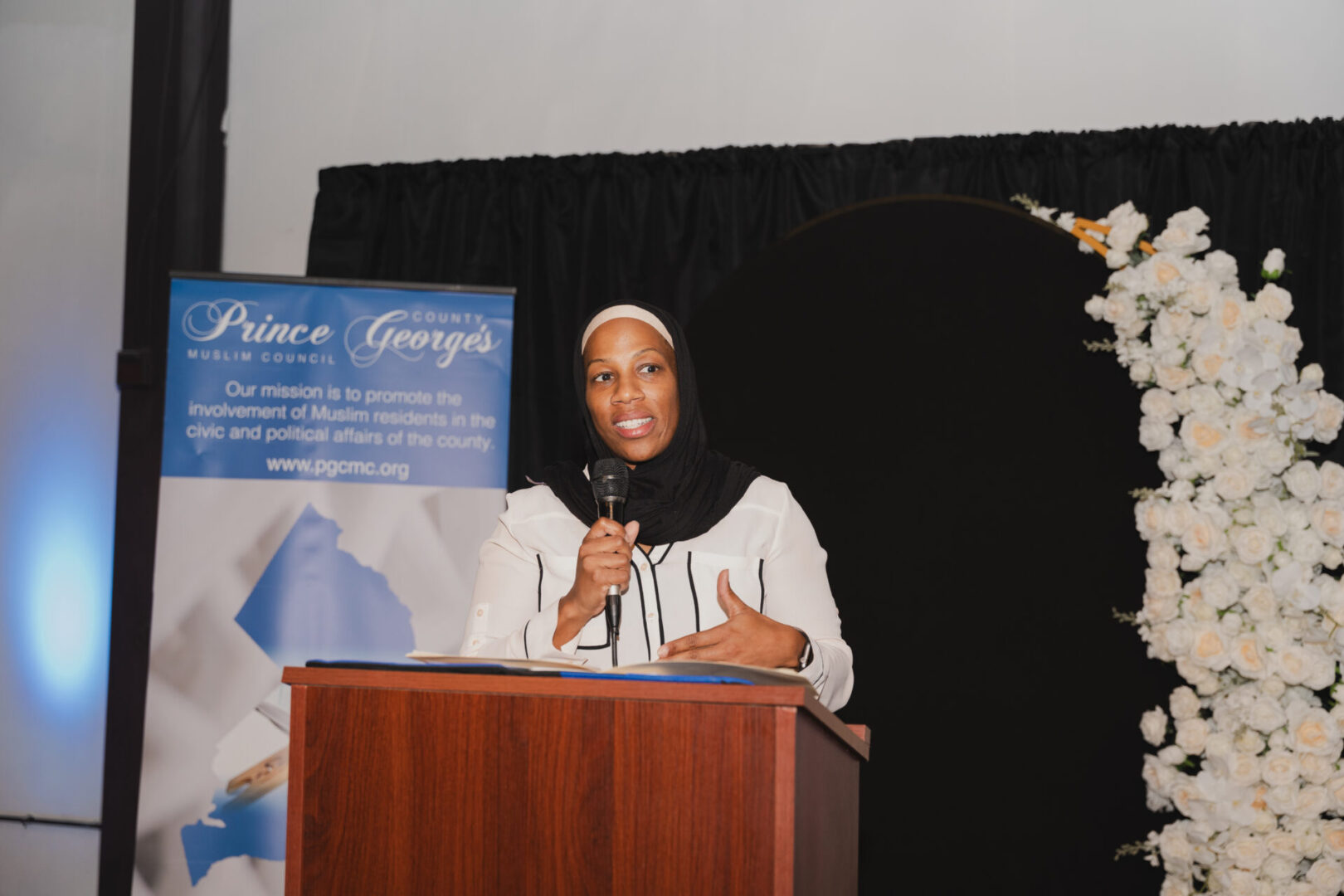 A woman speaks at a podium with a sign reading "Prince George's Muslim Council" and a floral backdrop on the right.