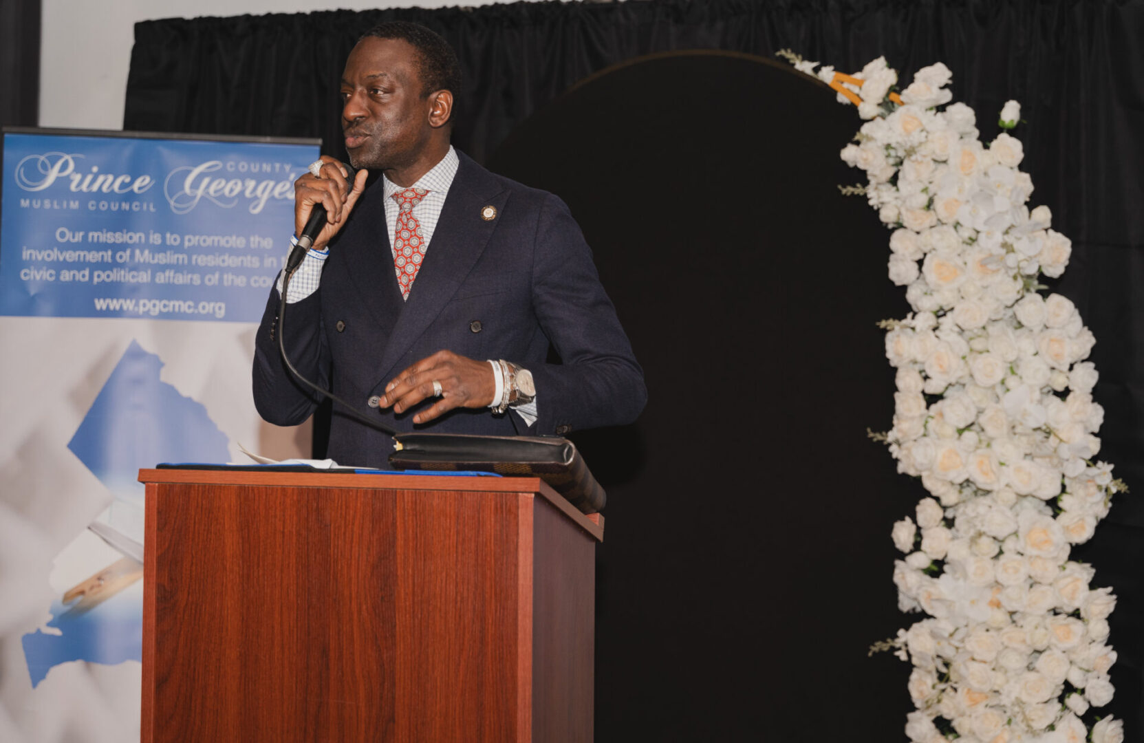 A man in a suit speaks at a podium with a microphone. A banner and flower arrangement are in the background.