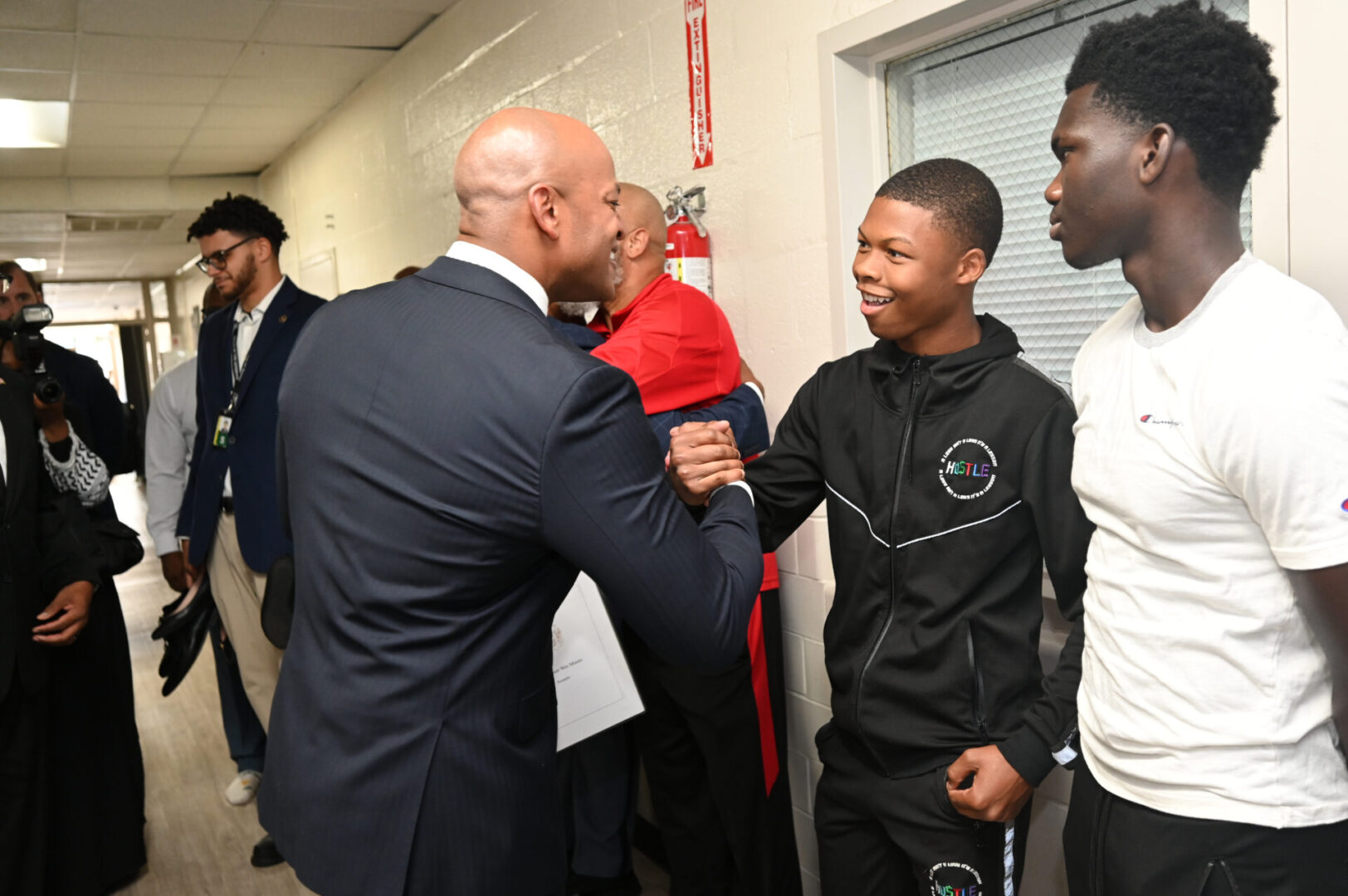 A man in a suit shakes hands with a young man in a tracksuit. Another young man stands nearby, with a few other people in the background.