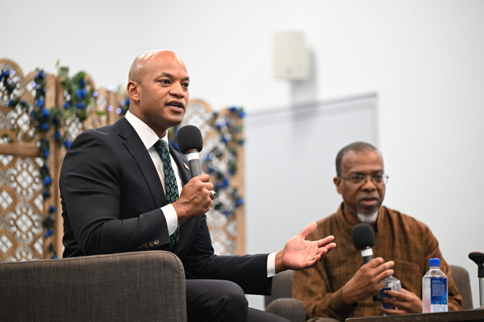 Two men seated, holding microphones. The man on the left wears a suit and gestures while speaking. The man on the right, in a brown shirt, holds a water bottle. Decorative panel in the background.