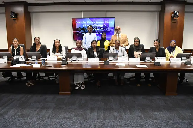 A group of people sit and stand around a conference table in a meeting room, with nameplates and laptops in front of them. A presentation screen is visible in the background.
