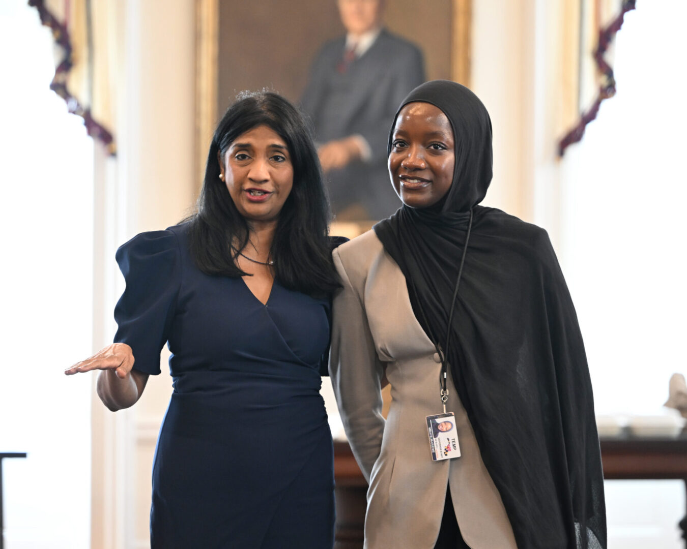 Two women stand side by side indoors; one gestures with her hand while the other smiles. A framed portrait is visible in the background.