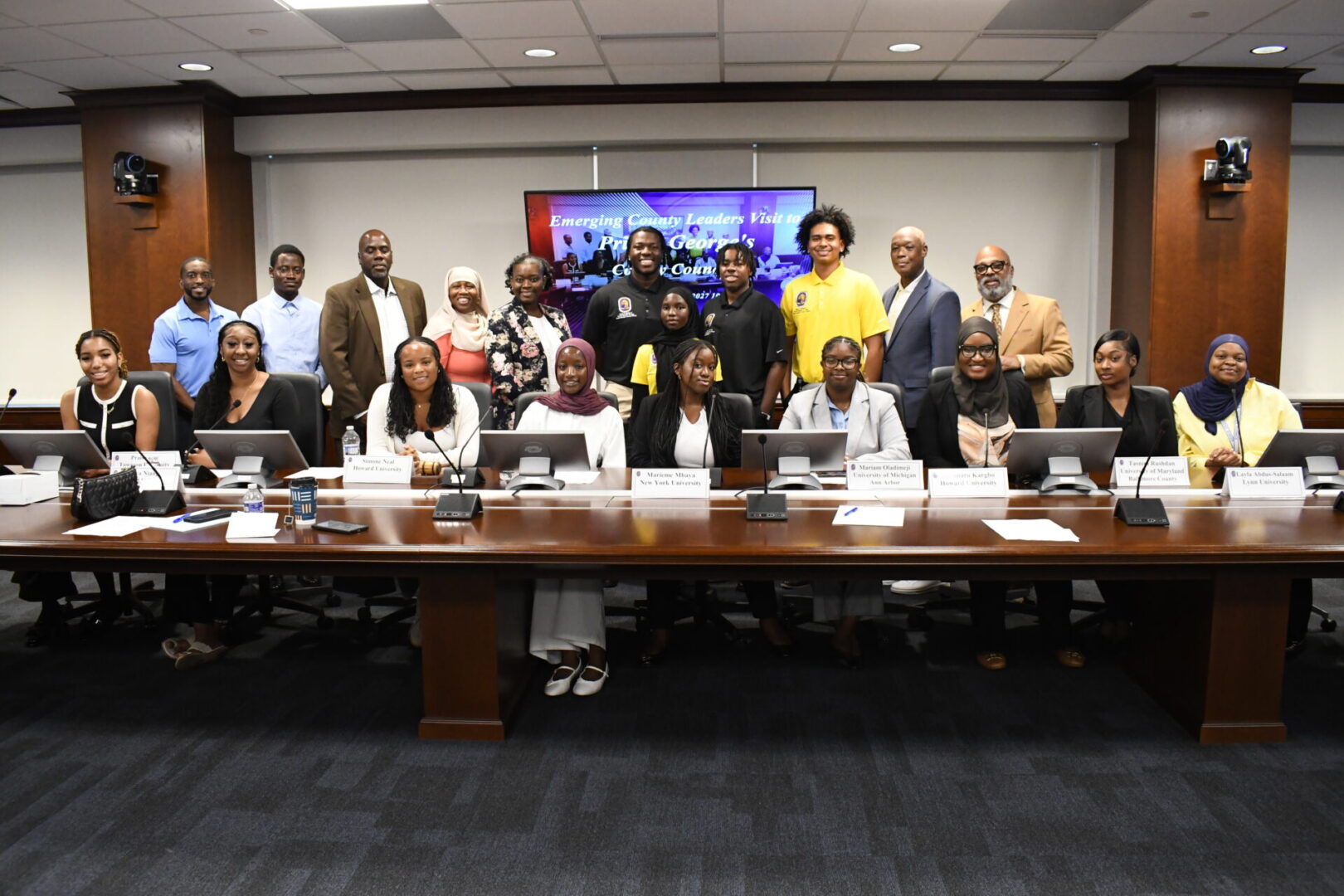 A group of people pose together in a conference room, with some seated at a long table and others standing behind them. A presentation screen is visible in the background.