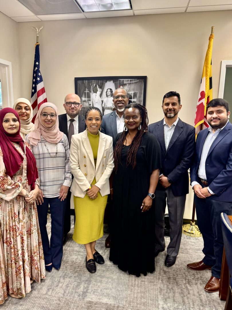 A group of nine people pose together in an office, standing in front of two flags and a framed photograph.