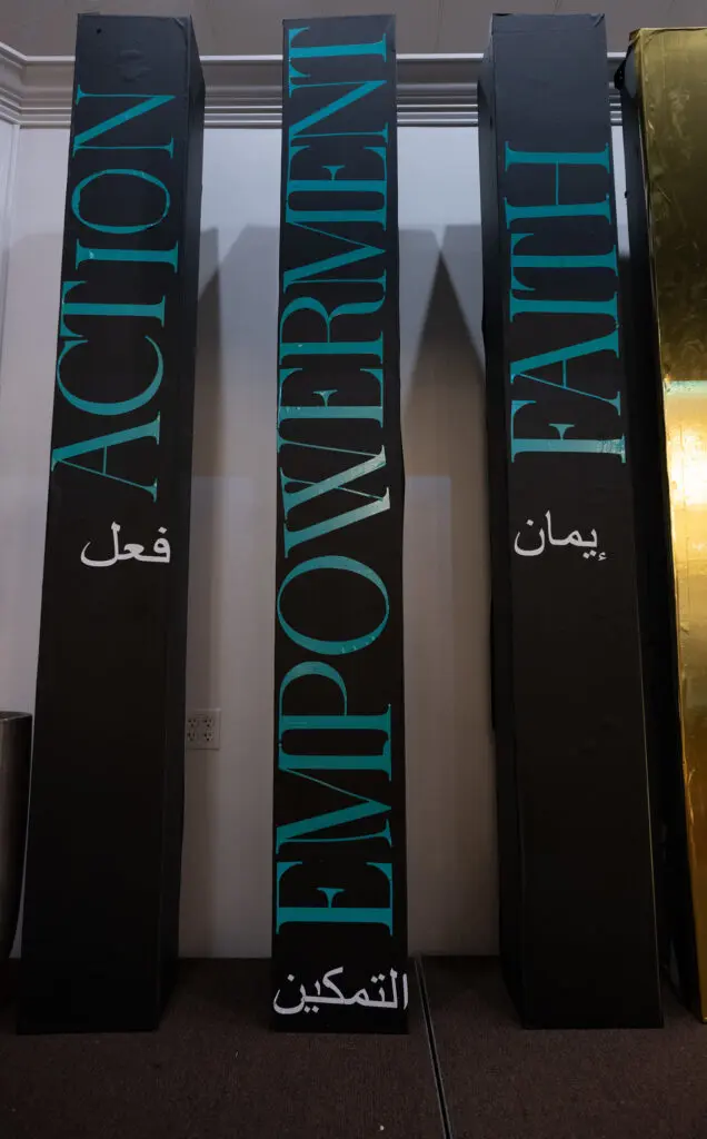 Three tall black pillars labeled "ACTION," "EMPOWERMENT," and "FAITH" in English and Arabic text, standing side by side indoors.