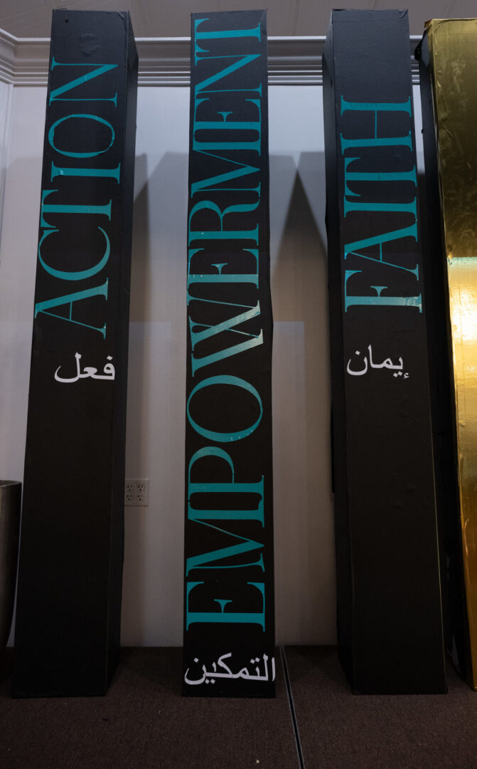 Three tall black pillars labeled "ACTION," "EMPOWERMENT," and "FAITH" in English and Arabic text, standing side by side indoors.