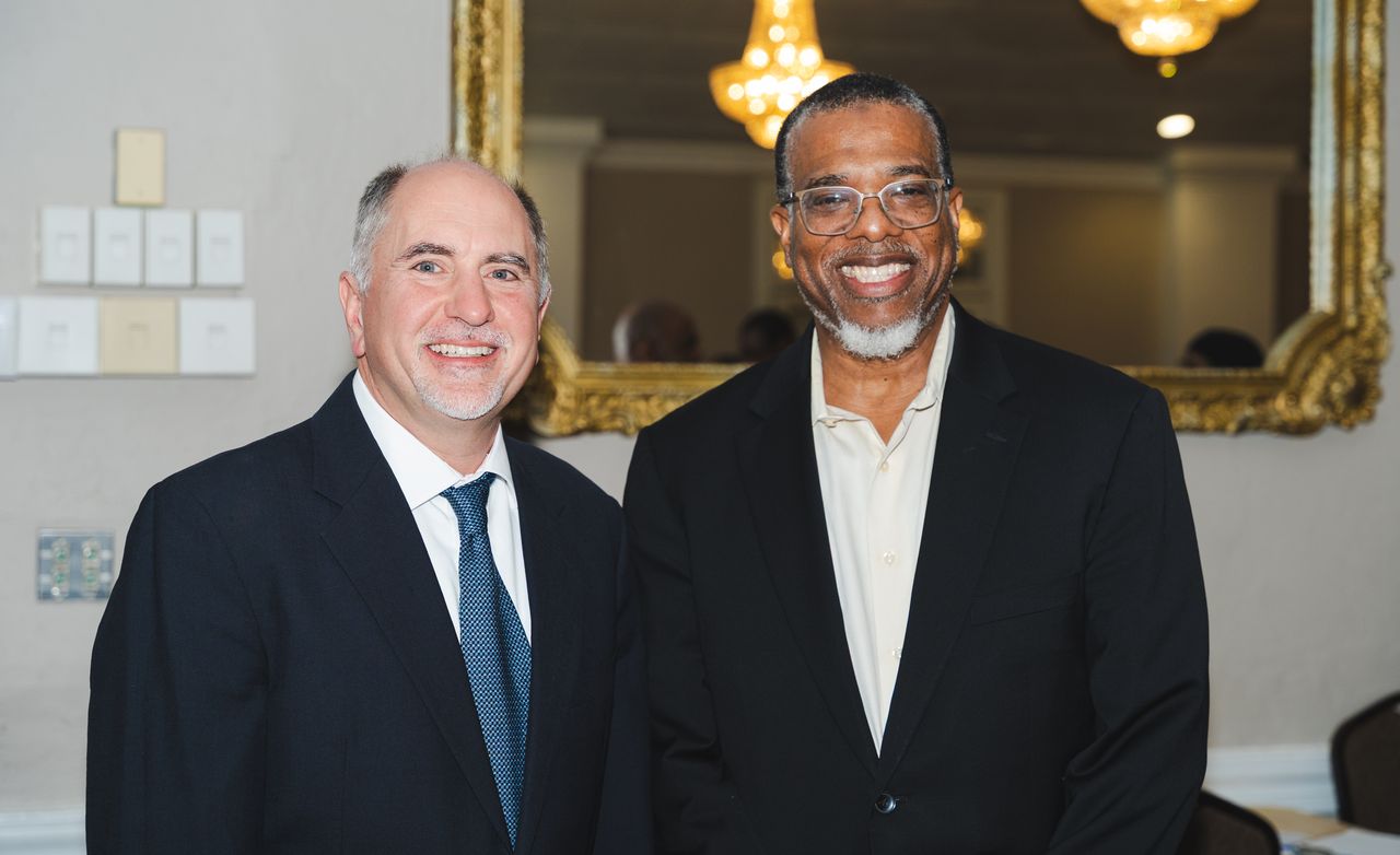Two men in suits stand side by side, smiling, in front of a large ornate mirror and chandeliers in a formal indoor setting.
