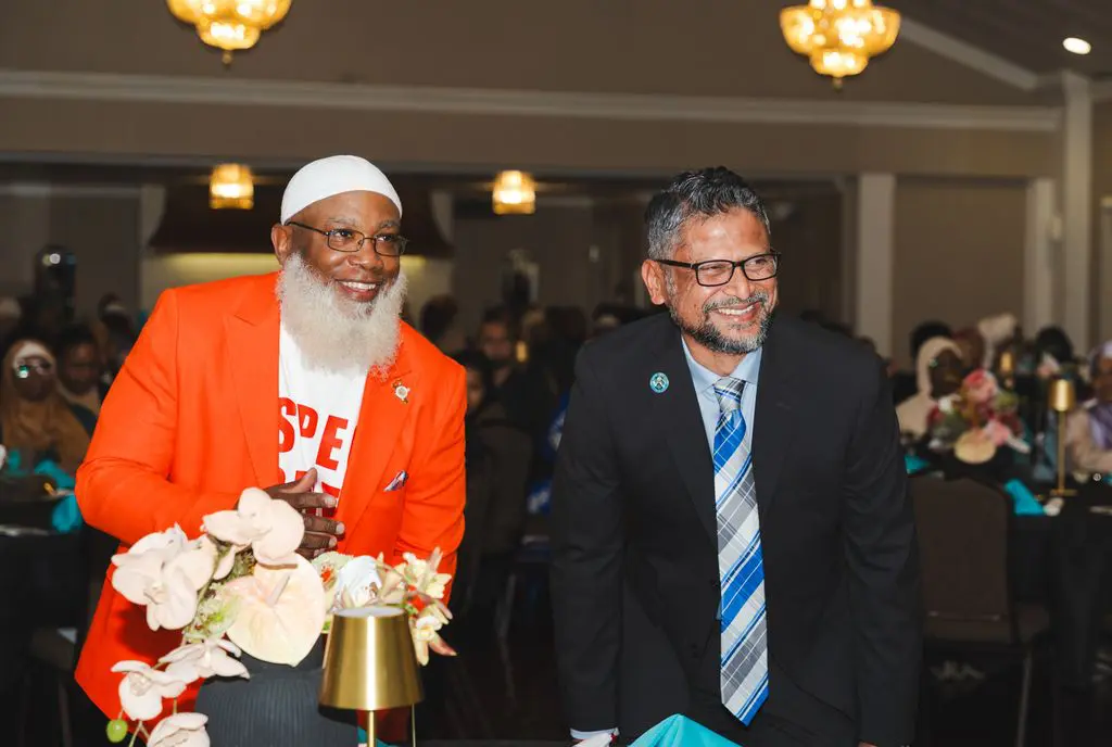 Two men in suits smile while standing next to each other at a formal indoor event with floral decorations and chandeliers.