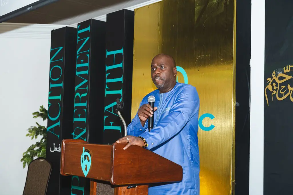 A man in a blue outfit speaks at a podium with the words "ACTION," "EXPERIMENT," and "FAITH" displayed vertically on banners behind him.