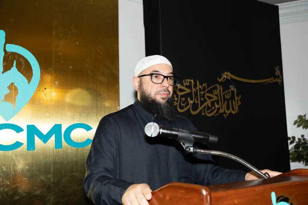 A man wearing glasses and a white cap speaks at a podium with a microphone. Arabic calligraphy and the letters "CMC" are visible in the background.